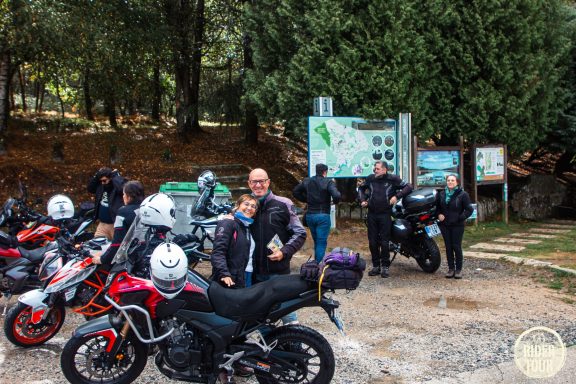 Grupo de motociclistas frente a un mapa en un área natural, con motos aparcadas.