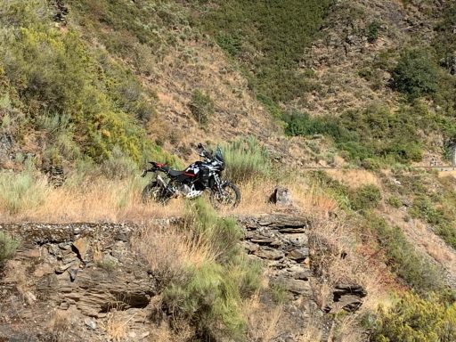 Motocicleta negra estacionada en un sendero montañoso y rocoso, rodeada de vegetación.